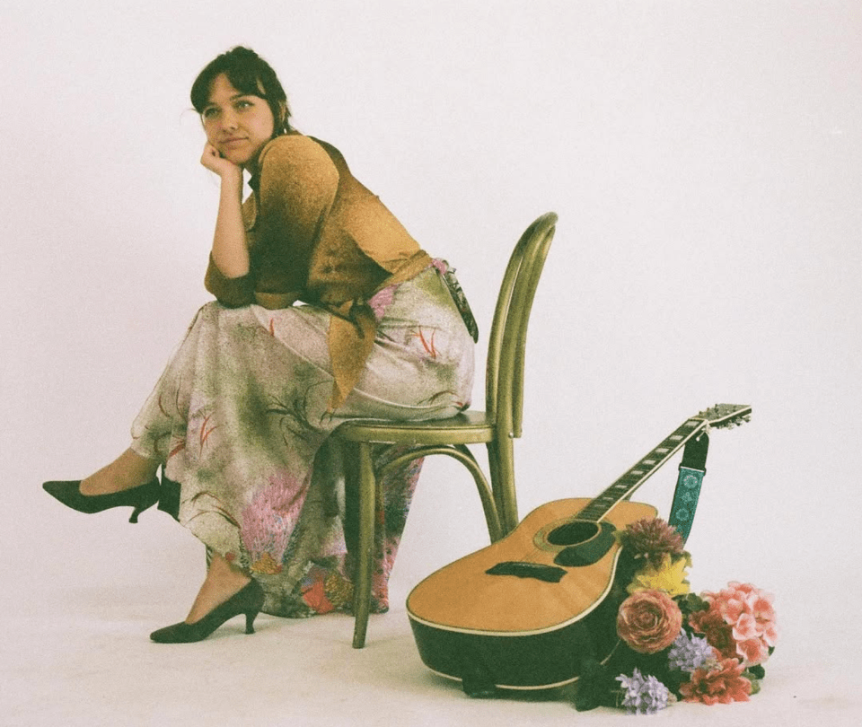 Arielle Soucy poses in a chair. She wears a floral skirt, and a guitar rests over a bouquet of flowers on the floor beside her.